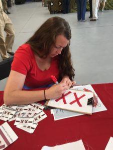 woman with long brown hair wearing a red shirt makes a quilt block of 2 red X's on a white base