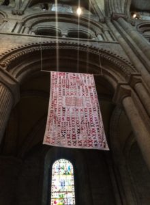 A quilt with a white background covered in pairs of red X’s hanging high in an ancient cathedral