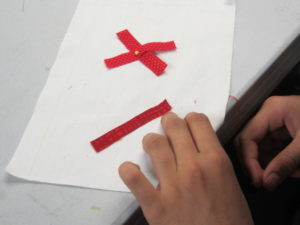 hands stitching a second red x onto a white piece of fabric