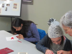 three people sitting at a table using needle and thread to stitch red strips of fabric onto white fabric