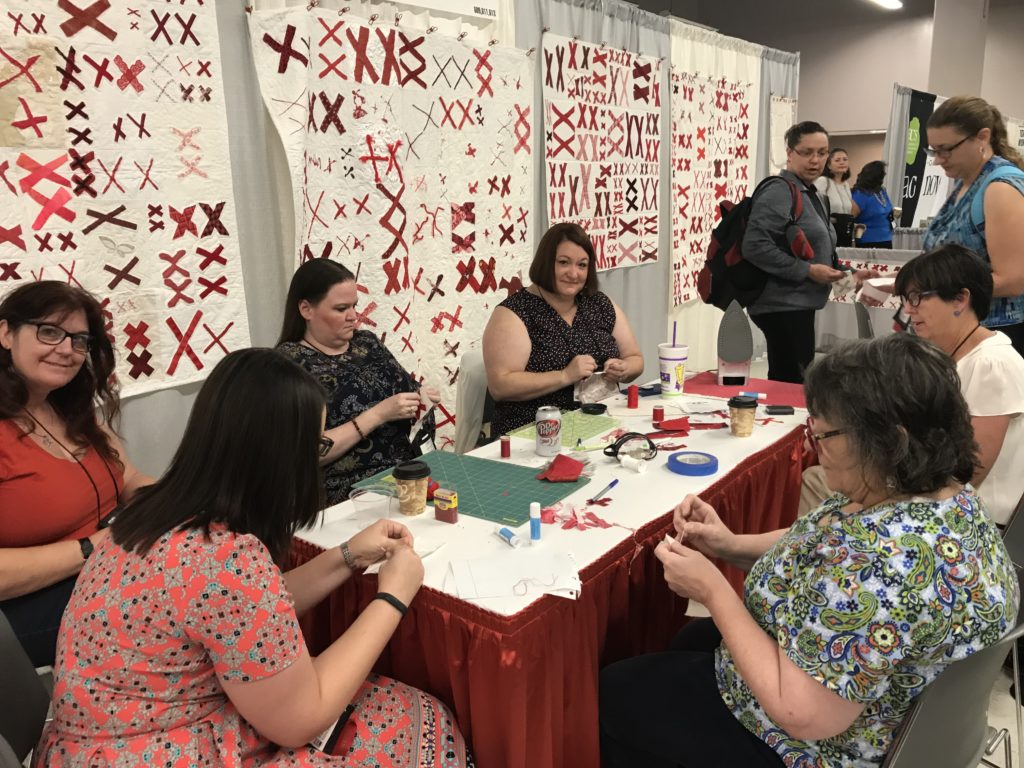 people sewing around a table with quilts hanging in the background